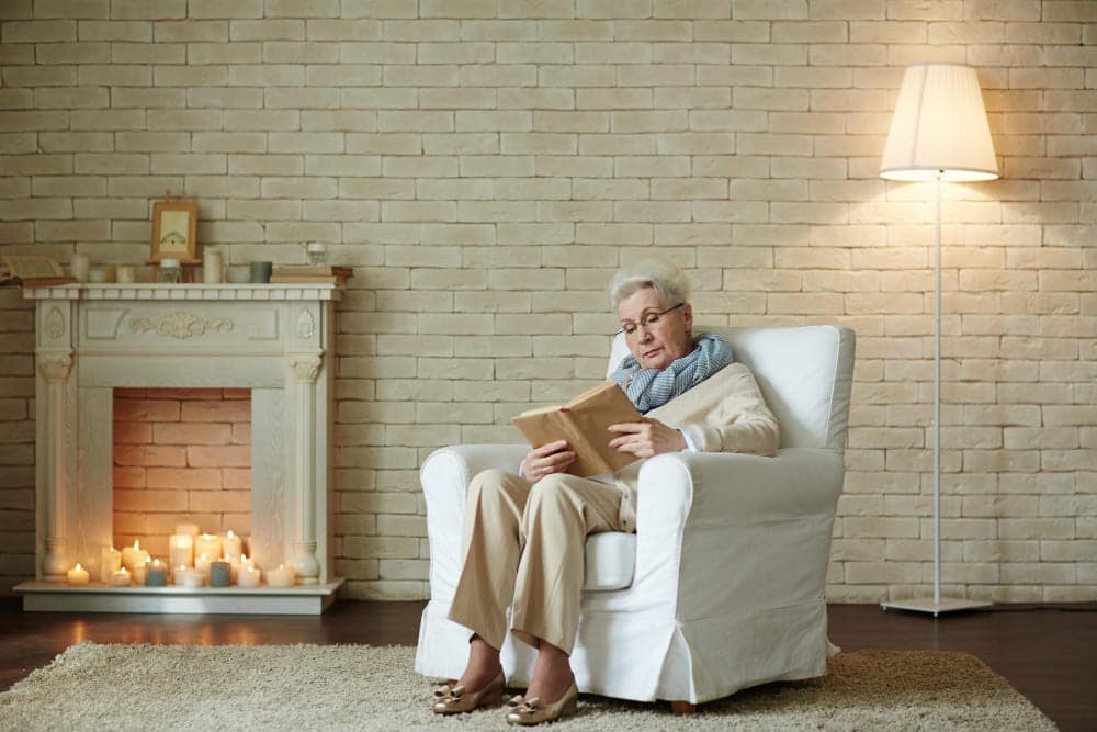 mujer mayor leyendo libro sentada en sillon frente a chimenea encendida en salon acogedor