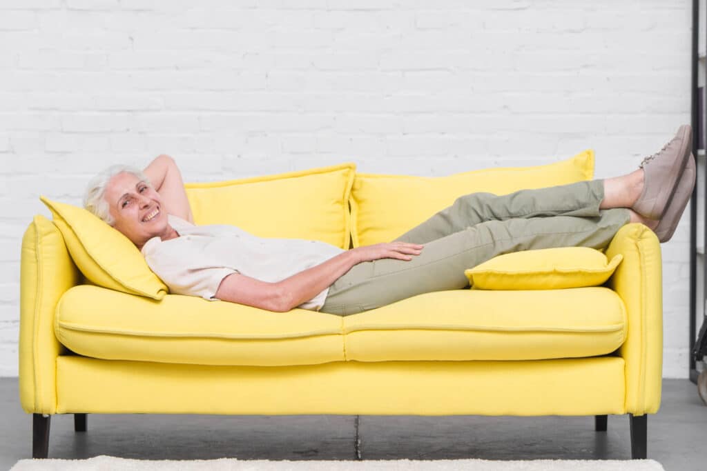 mujer mayor relajada tumbada en sofa amarillo en habitacion con pared blanca